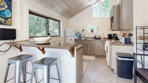 A kitchen with a central island, bar stools, a refrigerator, and a window overlooking greenery. - Geodesic Dome w/ Riverfront Deck, Lovely Views, & High-Speed WiFi - Dogs Welcome (Boothbay)