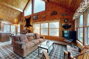 Ein gemütliches Blockhäuschen-Interieur mit kariertem Boden, einer Couch, einem Couchtisch, einem Fernseher und einem großen Fenster.