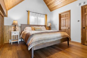 A bedroom with a wooden ceiling, a bed with a striped bedspread, two bedside lamps, a nightstand, and a door. - Lakefront Chalet-Style Home with Large Deck, a Dock & Beautiful Lake Views (Greenville)