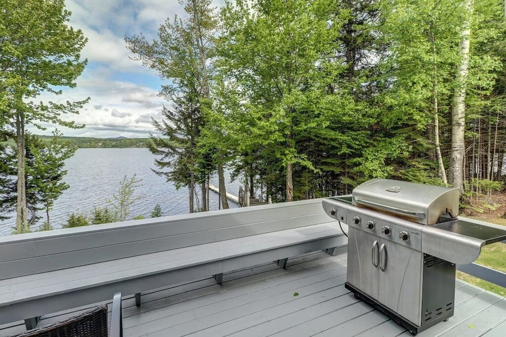 A deck with a grill overlooking a lake surrounded by trees.