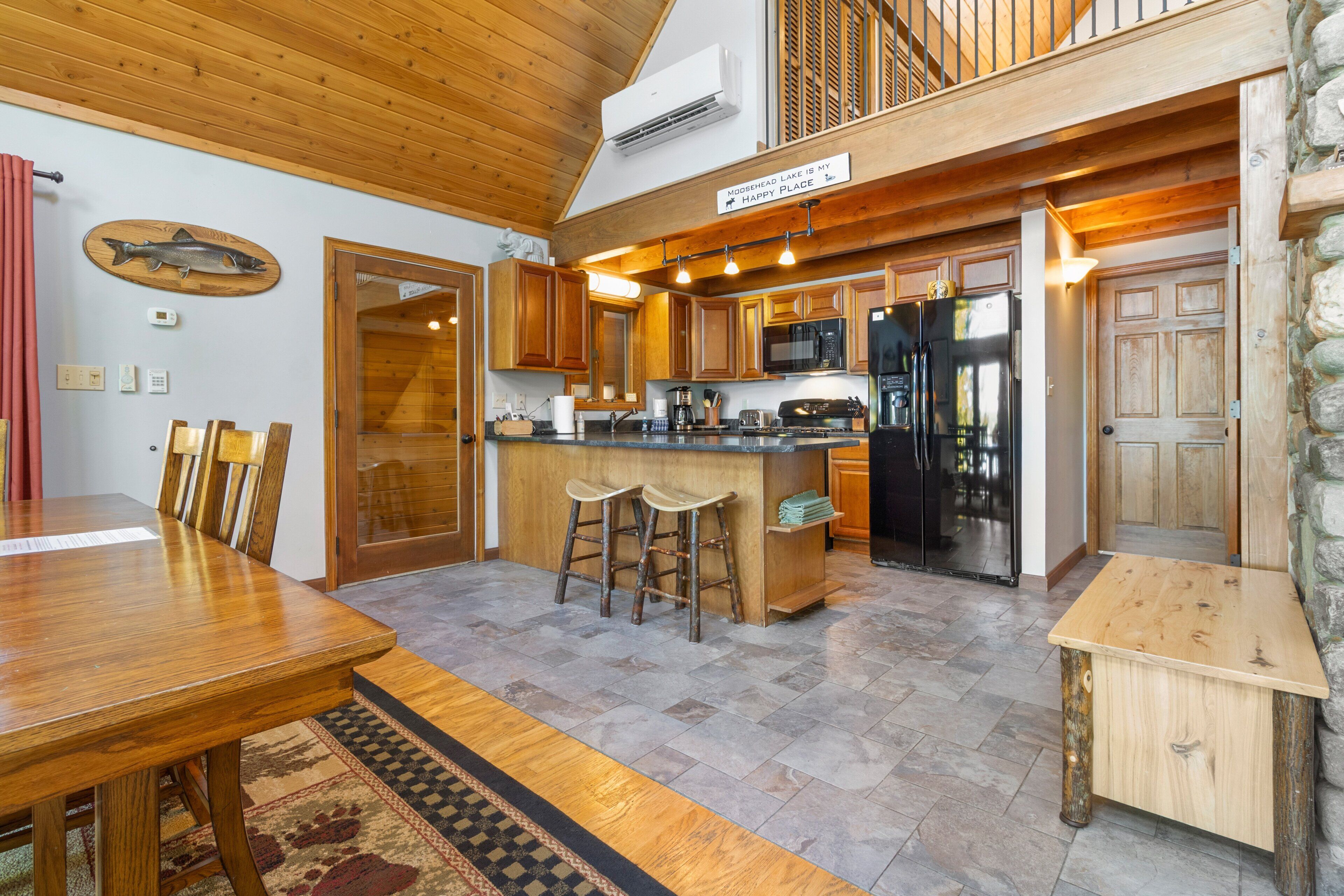 A spacious kitchen with wooden cabinets, a central island with stools, a black refrigerator, and a sign above the island.