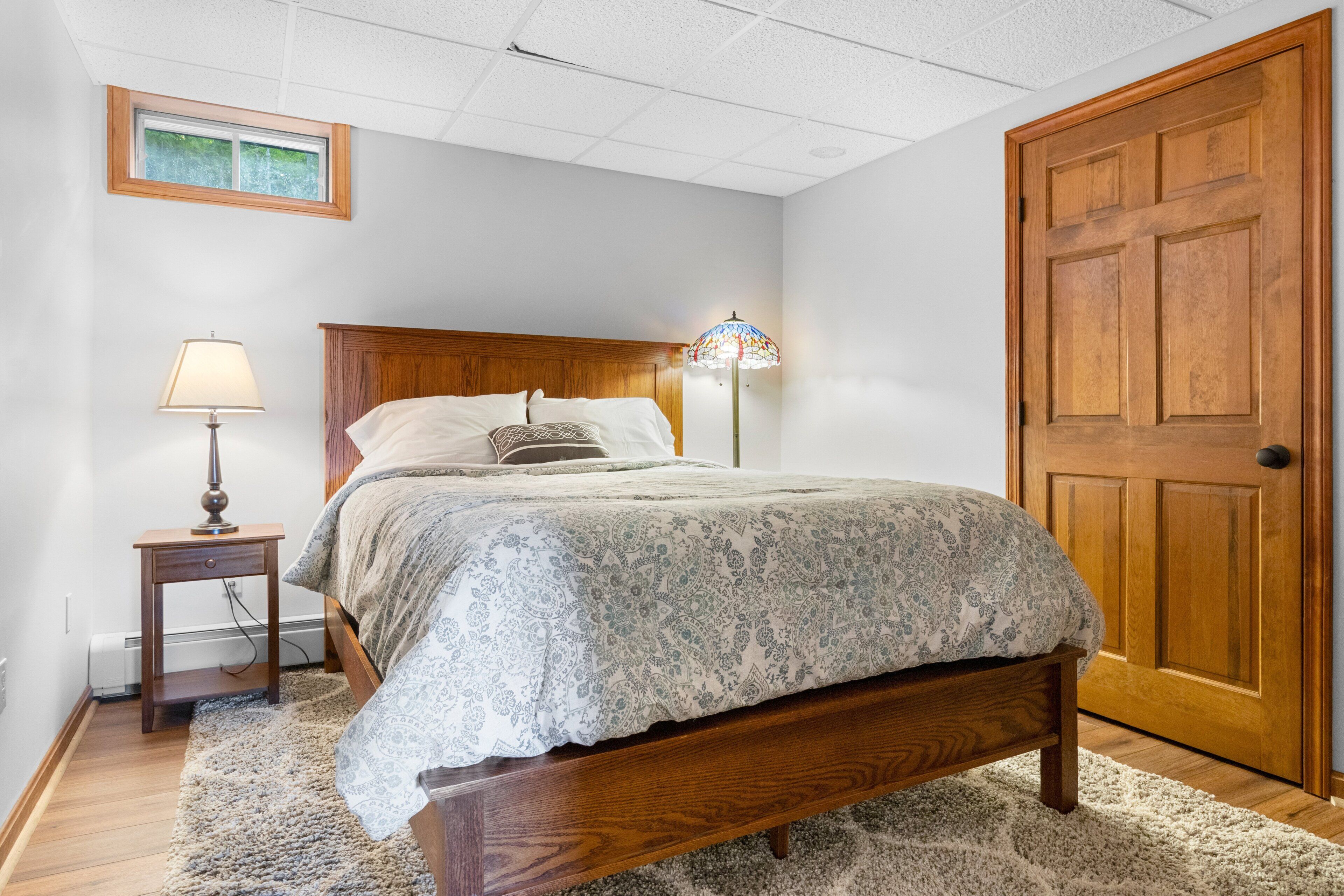 A bedroom with a wooden bed, a nightstand with a lamp, a floor lamp, a door, and a small picture on the wall.