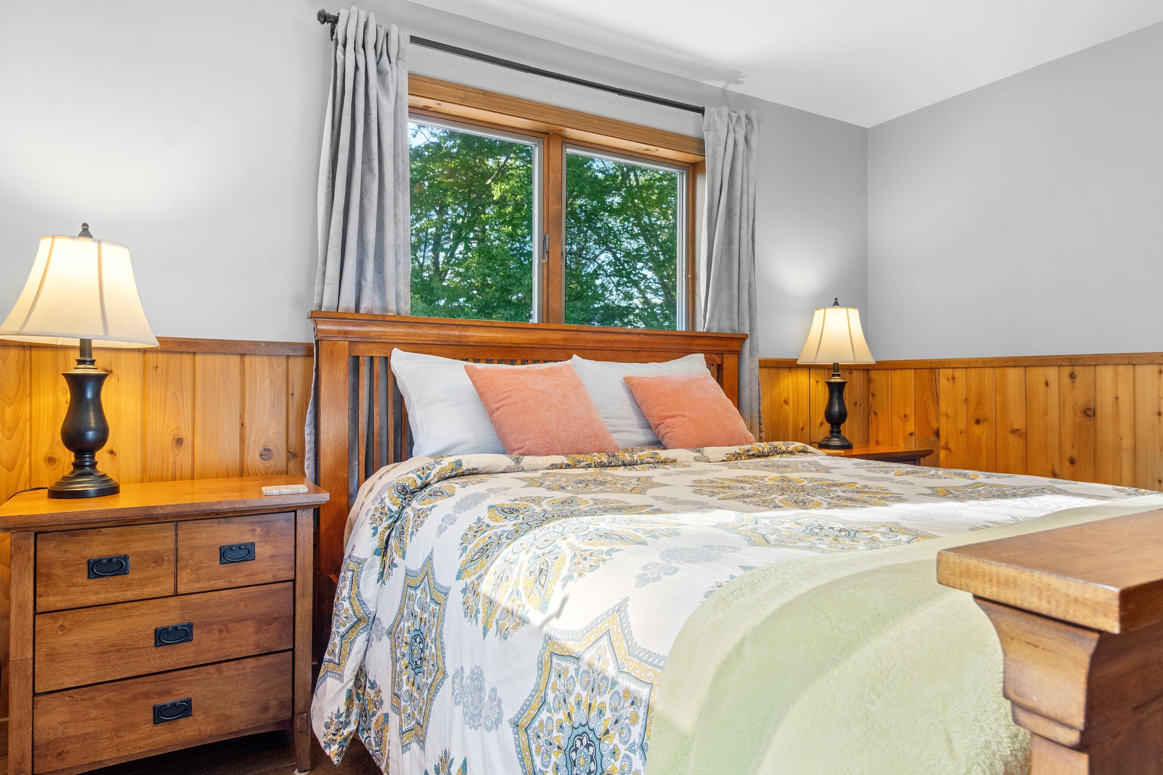 A bedroom with a wooden headboard, a bed with patterned bedding, two bedside lamps, a nightstand with drawers, and a window with curtains.