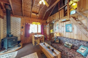 A rustic cabin interior with a brick wall, wooden furniture, a sofa with animal-themed cushions, a table with decorative items, and a staircase. - Lakefront cabin with private boat ramp, lake access, wrap-around porch, firepit (Lily Bay Township)