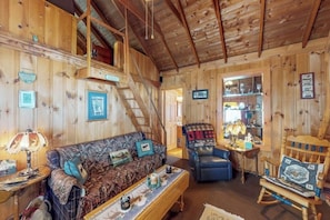 A wooden cabin interior with a sofa, armchair, and table, a staircase, and shelves with various items. - Lakefront cabin with private boat ramp, lake access, wrap-around porch, firepit (Lily Bay Township)
