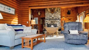 Interior de una cabaña de troncos con sofá, sillón, mesa de centro de madera, chimenea de piedra y trofeos de animales colgados.