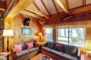 Interior de una cabaña con paredes de madera, un sofá, una mesa y un ventanal con vistas al exterior.