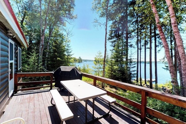 A deck with a table and grill, overlooking a lake and forest.