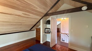 Een kamer met een schuine hemel, houten balken, een bed, een dressoir, een ingelijste foto en een trap die naar een ander niveau leidt.