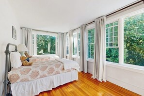 A bedroom with a large bed, a nightstand, a lamp, and a teddy bear. There are multiple windows with curtains.