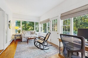 A room with a rocking chair, a desk with a computer, a sofa, and large windows.
