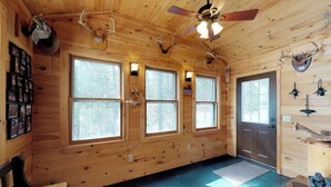 A cabin interior with wooden walls, large windows, a ceiling fan, and mounted antlers.