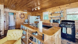 A wooden cabin interior with a kitchen area, a dining table, and a yellow armchair.