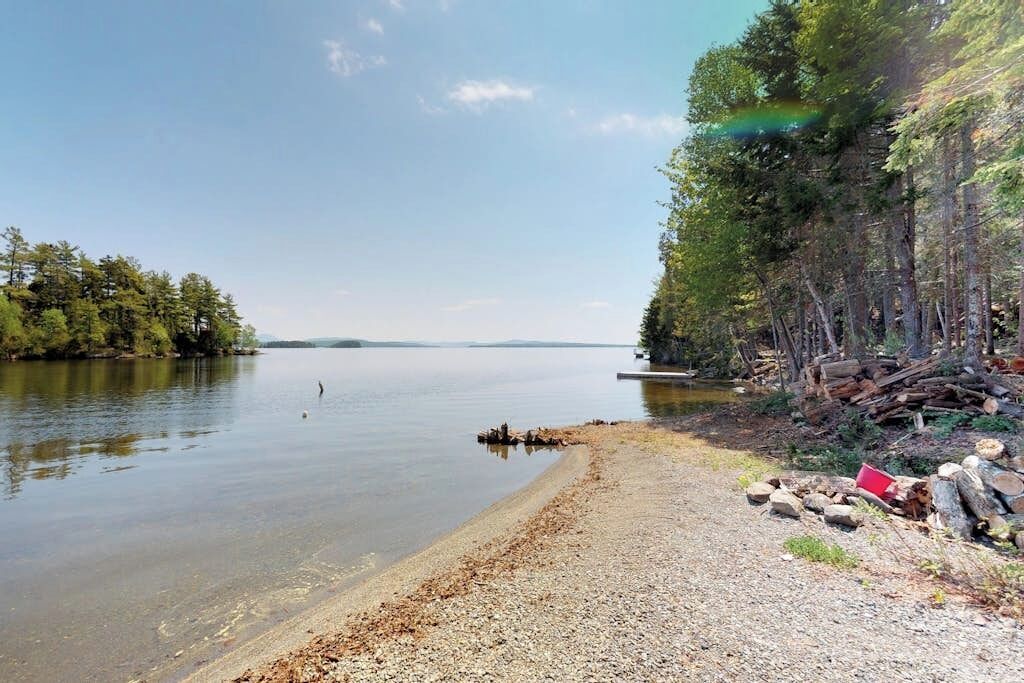 Un lac paisible aux rives de galets, bordé d’arbres et s’étendant jusqu’à une côte lointaine.