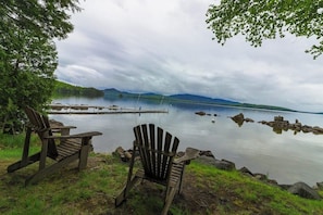 Two Adirondack chairs by a calm lake with a dock and mountains in the background.