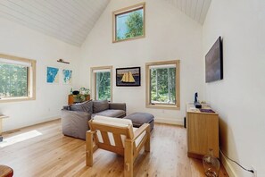 Ein geräumiger Raum mit hoher Decke, großen Fenstern, einem Holzstuhl, einer Couch und einem kleinen Schrank.
