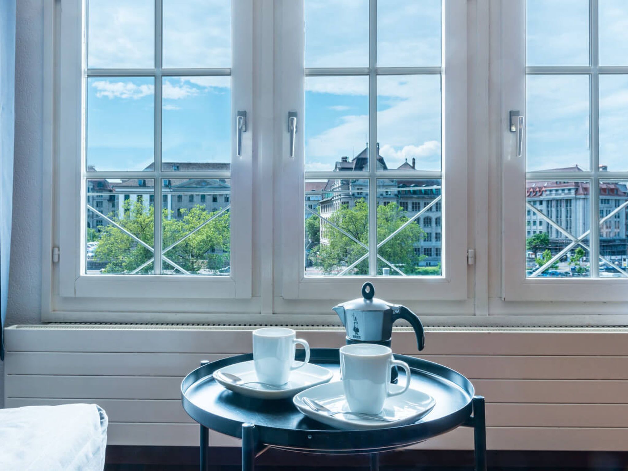 A room with a large window overlooking a cityscape, a small table with cups and a teapot, and a bed in the corner.