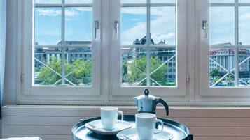 Ein Zimmer mit großem Fenster und Blick auf die Stadtlandschaft, ein kleiner Tisch mit Tassen und einem Teekessel sowie ein Bett in der Ecke.