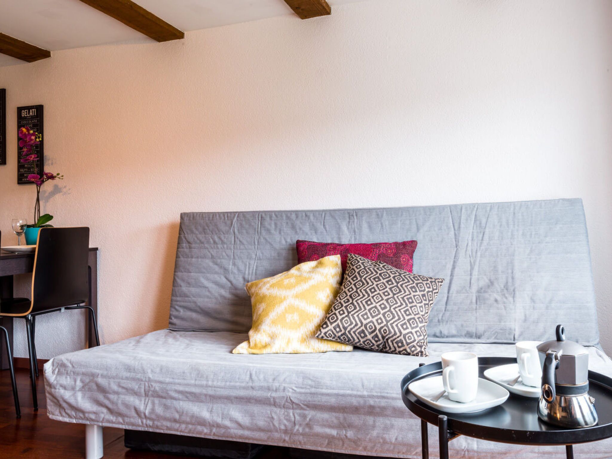 Una habitación moderna y minimalista con un sofá gris, dos almohadas estampadas, una mesita redonda con cafetera y una silla.