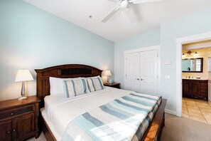 Une chambre à coucher avec un lit, des tables de chevet, un ventilateur de plafond, une porte et une salle de bain visible à travers une porte ouverte.