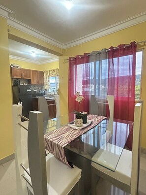 A dining area with a glass table, chairs, a kitchen in the background, and a window with curtains. - 2-bedrooms apartment in peaceful Concepción La Vega with AC (Concepción La Vega)