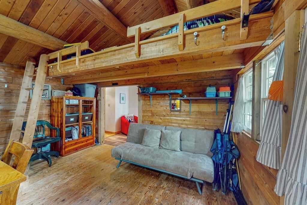 A cozy cabin interior with a loft bed, a sofa, a wooden bookshelf, and a window with curtains.