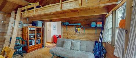 A cozy cabin interior with a loft bed, a sofa, a wooden bookshelf, and a window with curtains.