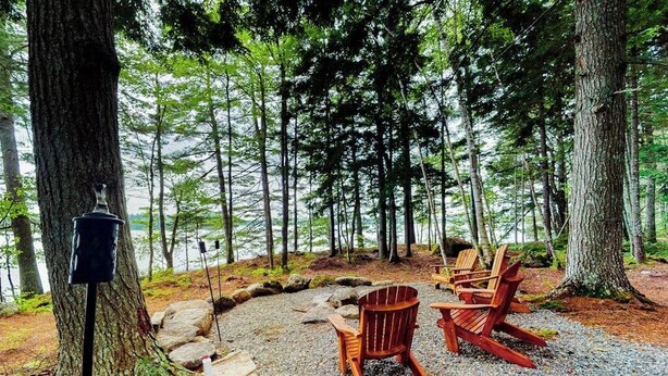 Een bebost gebied met een grindpad, houten Adirondack-stoelen en een vogelvoeder.