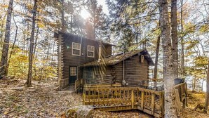 A log cabin with a deck, surrounded by trees and a forest floor covered with fallen leaves. - 2 beautifully designed, off-the-grid homes with amazing lakefront views & access (Franklin)