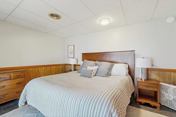 Une chambre avec un grand lit, une tête de lit en bois, des tables de chevet avec des lampes, un ventilateur et un tableau encadré au mur.