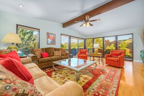 Ein geräumiges Wohnzimmer mit hoher Decke, großen Fenstern mit Blick ins Freie, einem Deckenventilator und verschiedenen Sitzmöglichkeiten, darunter ein Sofa und zwei Sessel.
