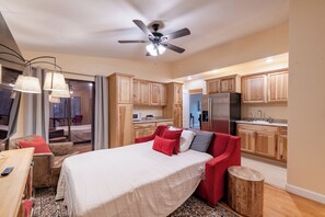 Ein Schlafzimmer mit einem Bett, einem roten Sessel, einem hölzernen Hocker und einer Küchenzeile mit Holzschränken und einem Kühlschrank.