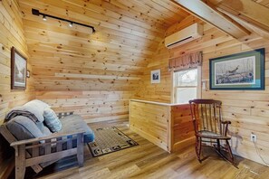 A wooden cabin interior with a bed, a rocking chair, a bench, and a window with a view.