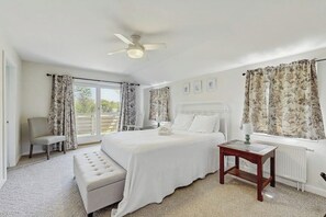 Un dormitorio con cama, un banco, una mesa, un ventilador de techo y una ventana con cortinas.
