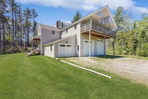 A two-story house with a covered porch, a garage, and a large lawn. - Modern farmhouse with deck, firepit, & balcony (Northport)