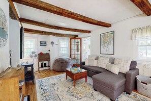 A cozy living room with a sofa, a coffee table, a wooden desk, and a large window. - Charming Boothbay Harbor cottage with partial AC, full kitchen, and sun room (Boothbay Harbor)