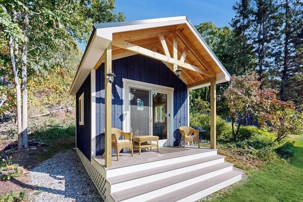 A small cabin with a porch, two wicker chairs, and a wooden deck. - Charming Boothbay Harbor cottage with partial AC, full kitchen, and sun room (Boothbay Harbor)