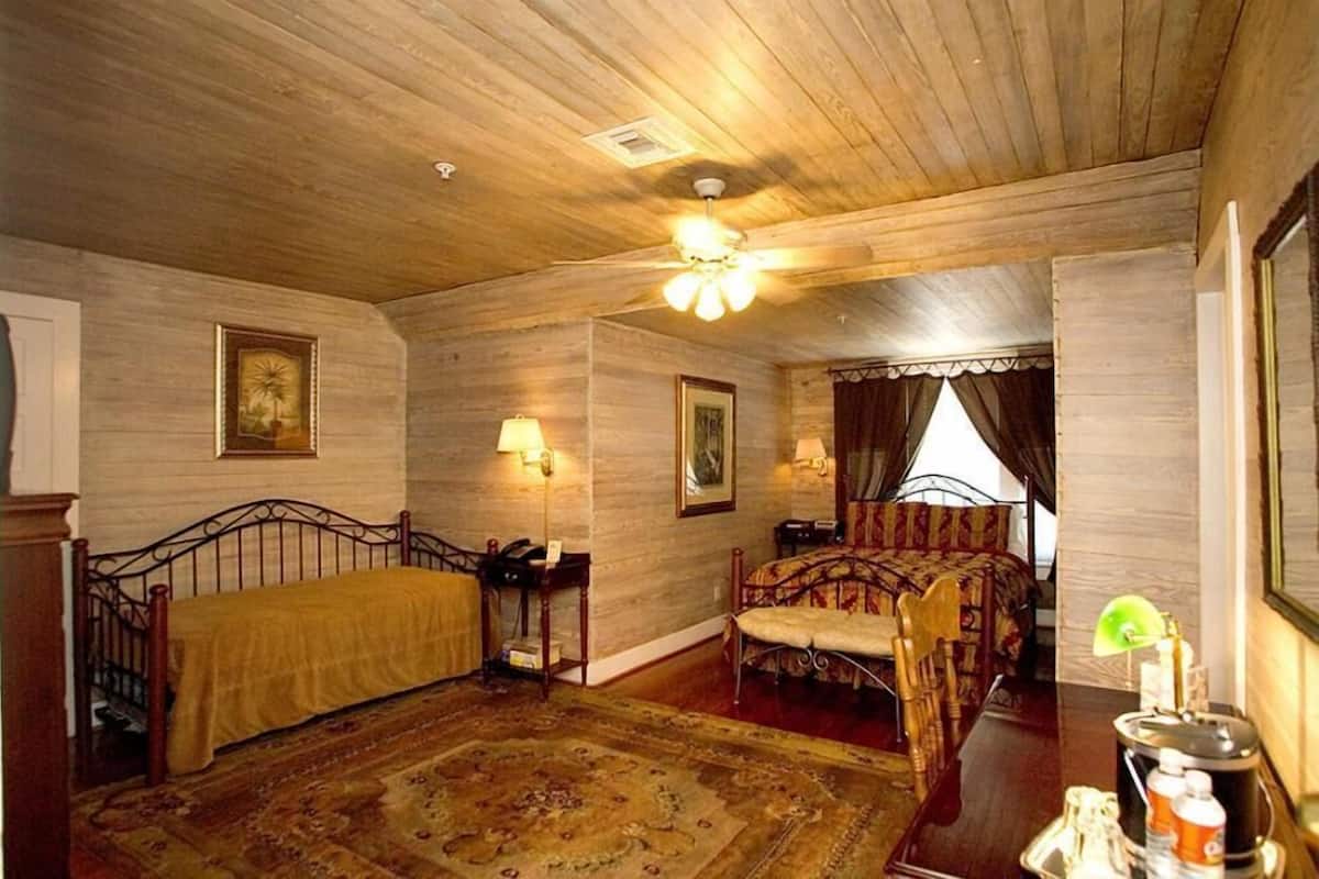 Una habitación con techo de madera, una cama con colcha estampada, una mesita de noche con lámpara y una ventana con cortinas.