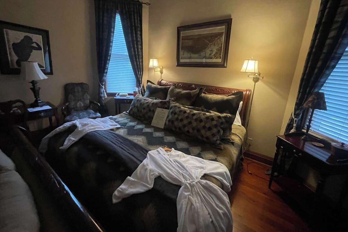 Una habitación de hotel con una cama, dos lámparas de noche, una silla, un escritorio y una ventana con cortinas.