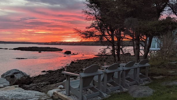 A sunset view with a rocky shoreline, wooden chairs, and a distant tree line. - Ocean Point Cottage- large porch, ocean view, water access (East Boothbay)