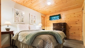 Une chambre avec un plafond en bois, un lit avec une literie à carreaux, deux lampes de chevet, une commode et une fenêtre avec un rideau.