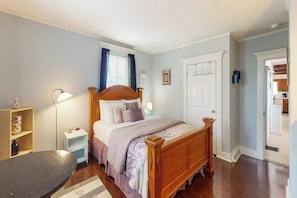 Quarto com cama de madeira, mesas de cabeceira, um criado-mudo, abajur e uma janela com cortinas.