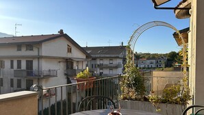 A round wooden table with a clear glass container and a white pitcher on a balcony with a metal railing, overlooking a residential area with houses and a clear sky.