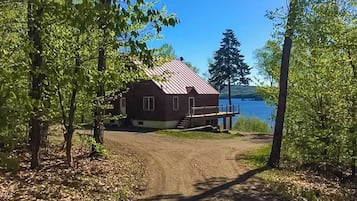 Une cabane avec un toit en métal, un chemin de terre menant à celle-ci, et un lac en arrière-plan.