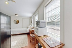 Een kleine kamer met een bed, een bureau met een computer, een stoel en een raam met jaloezieën.