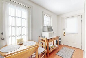 Ein Zimmer mit einem Holztisch, zwei Stühlen, einer Mikrowelle, einem kleinen Regal mit Büchern und einer Tür mit Fenster.