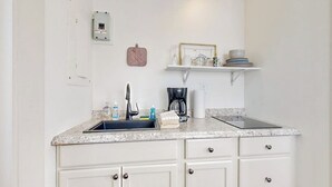 Una cocina con un lavamanos, encimera, armarios, cafetera y una repisa con vajilla.