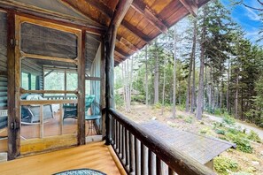 A wooden cabin with a deck, a covered porch, and a view of a forest.