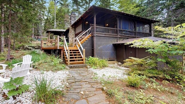 A cabin with a stone pathway leading to a wooden deck, surrounded by trees and ferns.