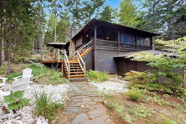 A cabin with a stone pathway leading to a wooden deck, surrounded by trees and ferns.